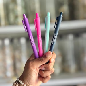 May include: A hand holds four pens in purple, pink, teal, and navy blue. Each pen has a different phrase printed on it: "THE BOOK WAS BETTER", "*ADDS TO TBR", "READ MORE BOOKS", and "JUST ONE MORE CHAPTER". The pens are a writing accessory.