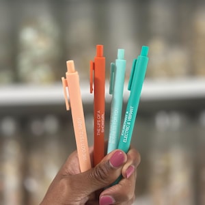 Peut inclure: Quatre stylos colorés, pêche, orange, bleu clair et turquoise, sont tenus dans une main. Chaque stylo porte une inscription différente : "WE GOT TS12, BABY", "THE LIFE OF A SHOWGIRL", "BUSINESS FOR YOU" et "EXUBERANT & ELECTRIC & VIBRANT".