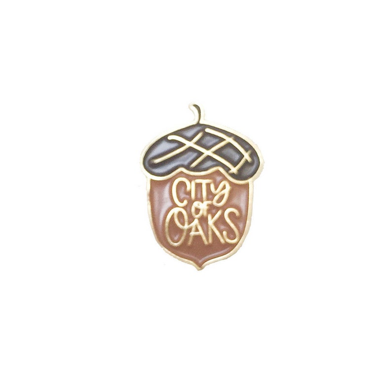 May include: A gold acorn pin with a brown and black enamel design. The acorn has the words "City of Oaks" written on it.