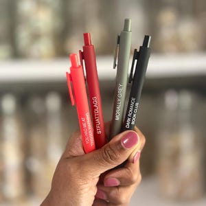 Può includere: Quattro penne rosse, rosso scuro, grigie e nere sono tenute in mano. La penna rossa recita "YOU HAD ME AT TRIGGER WARNING". La penna rosso scuro recita "STFUATTDLAGG". La penna grigia recita "MORALLY GREY". La penna nera recita "DARK ROMANCE BOOK CLUB."