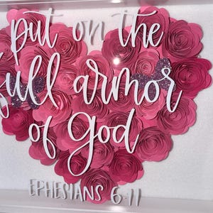 May include: A framed artwork featuring a heart-shaped arrangement of pink paper flowers. White script reads "put on the full armor of God" with "Ephesians 6:11" below. Two glittery purple butterflies accent the floral design.