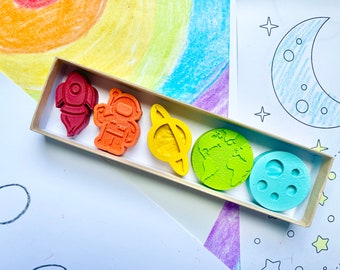 May include: A set of six colorful crayons shaped like space-themed objects, including a rocket, astronaut, planet, Earth, moon, and a ringed planet. The crayons are in a cardboard box.