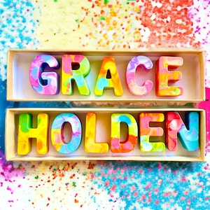 Op de afbeelding: Een set kleurrijke, gemarmerde waskrijtletters die de naam "GRACE HOLDEN" spellen. De letters zijn in twee rijen in een houten doos gerangschikt.