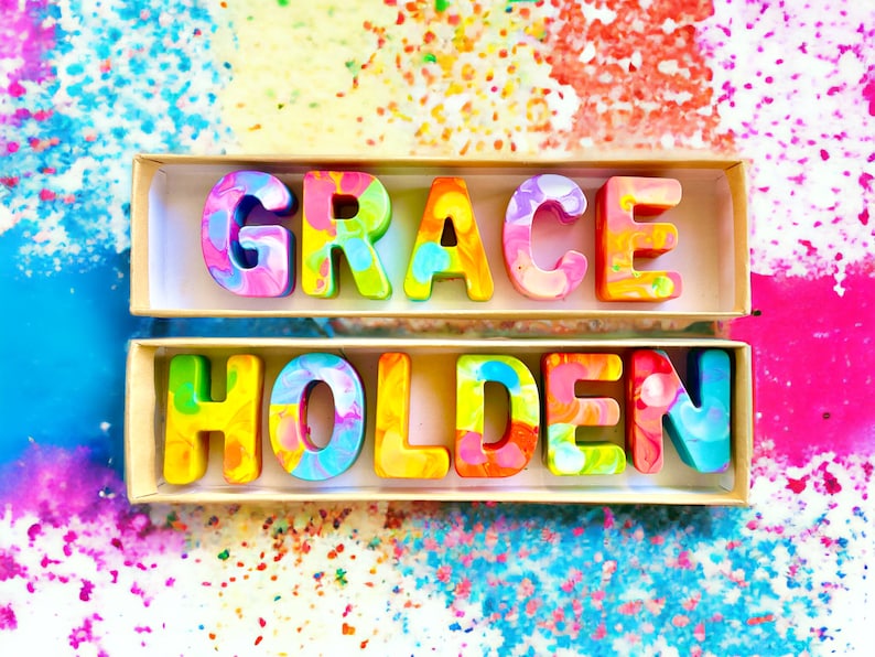 Op de afbeelding: Een set kleurrijke, gemarmerde waskrijtletters die de naam "GRACE HOLDEN" spellen. De letters zijn in twee rijen in een houten doos gerangschikt.