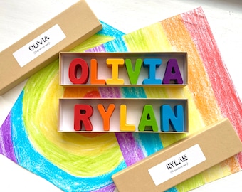May include: Two boxes with the names "Olivia" and "Rylan" printed on them. The boxes contain colorful wooden letters spelling out the names "Olivia" and "Rylan". The boxes are sitting on a rainbow-colored drawing.
