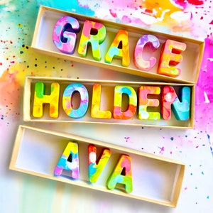 Op de afbeelding: Drie dozen met kleurrijke, gemarmerde letters die de namen "Grace", "Holden" en "Ava" spellen. De dozen staan op een witte achtergrond met kleurrijke verfspatten.