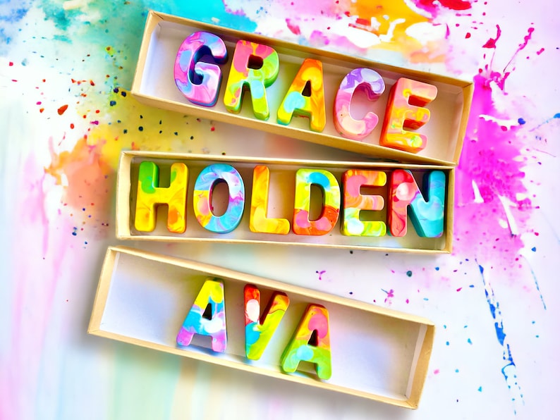 Op de afbeelding: Drie dozen met kleurrijke, gemarmerde letters die de namen "Grace", "Holden" en "Ava" spellen. De dozen staan op een witte achtergrond met kleurrijke verfspatten.