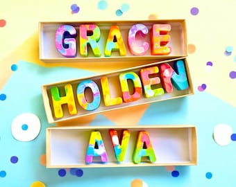 以下が含まれることがあります： 「GRACE」、「HOLDEN」、「AVA」という名前を綴る、カラフルで渦巻き状のクレヨン文字が入った3つの箱。