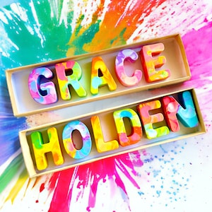 Op de afbeelding: Een houten doos met de naam "GRACE HOLDEN" gespeld in kleurrijke, gemarmerde letters. De letters zijn in twee regels gerangschikt, met "GRACE" op de bovenste regel en "HOLDEN" op de onderste regel. De doos staat op een wit oppervlak met een kleurrijke, abstracte achtergrond.