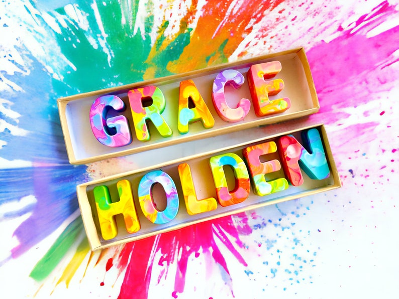 Op de afbeelding: Een houten doos met de naam "GRACE HOLDEN" gespeld in kleurrijke, gemarmerde letters. De letters zijn in twee regels gerangschikt, met "GRACE" op de bovenste regel en "HOLDEN" op de onderste regel. De doos staat op een wit oppervlak met een kleurrijke, abstracte achtergrond.