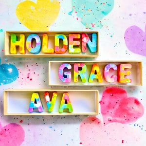 Op de afbeelding: Drie houten dozen met kleurrijke letters die de namen Holden, Grace en Ava spellen. De dozen staan op een witte achtergrond met roze, blauwe en gele harten.