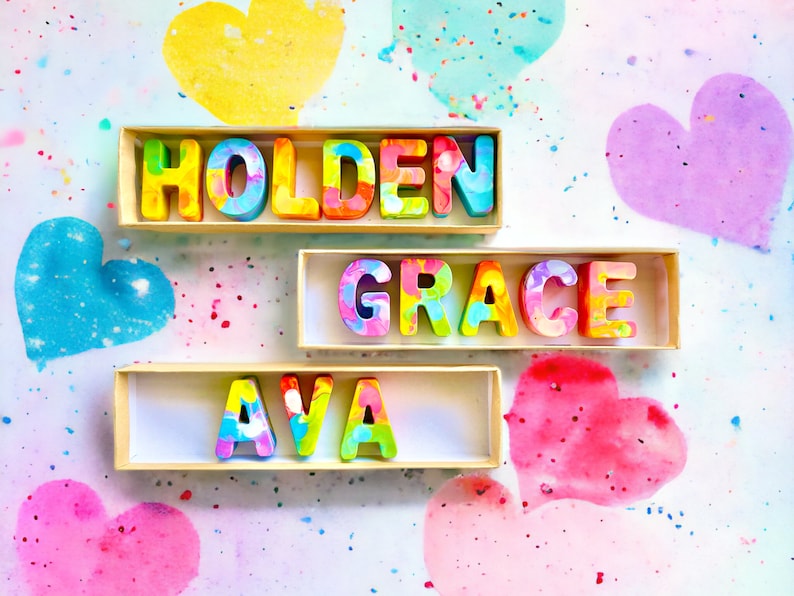 Op de afbeelding: Drie houten dozen met kleurrijke letters die de namen Holden, Grace en Ava spellen. De dozen staan op een witte achtergrond met roze, blauwe en gele harten.