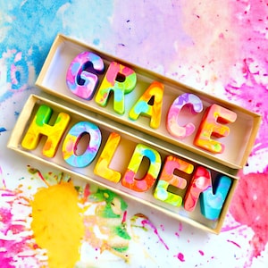 Op de afbeelding: Twee dozen gevuld met kleurrijke, gemarmerde, wasletters die de naam "GRACE HOLDEN" spellen. De letters bevinden zich in een lichtbruine doos op een witte achtergrond met kleurrijke waterverfsplatters.