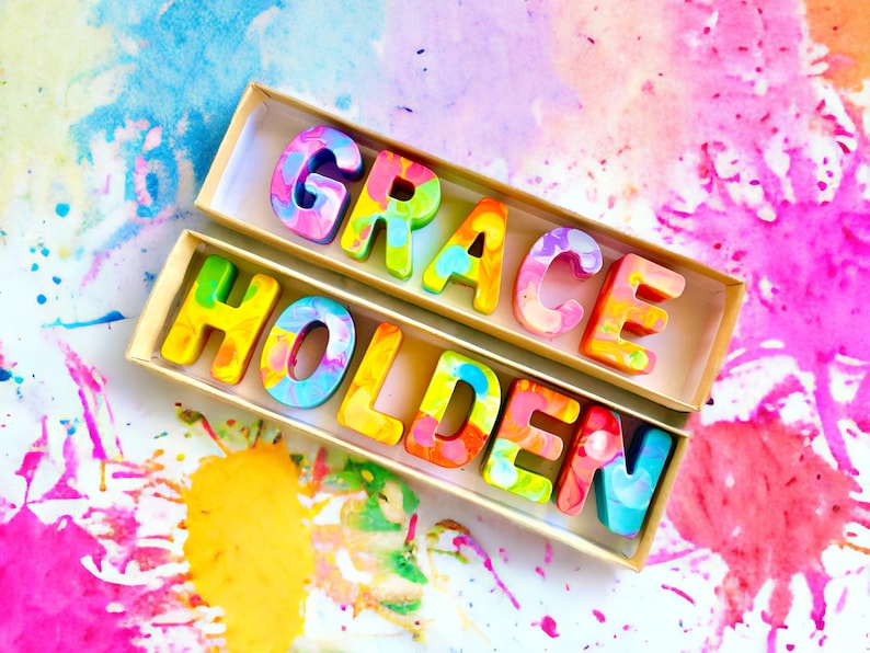 Op de afbeelding: Twee dozen gevuld met kleurrijke, gemarmerde, wasletters die de naam "GRACE HOLDEN" spellen. De letters bevinden zich in een lichtbruine doos op een witte achtergrond met kleurrijke waterverfsplatters.