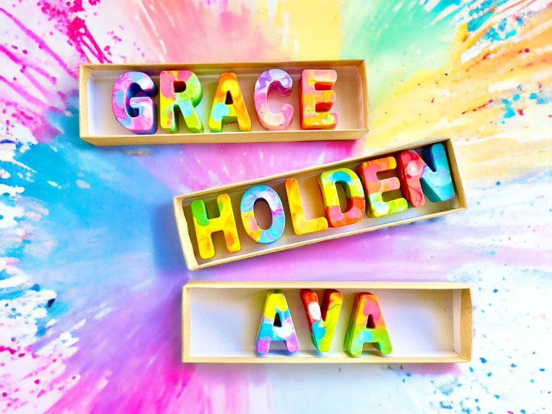 Op de afbeelding: Drie houten dozen met kleurrijke waskrijtletters die de namen "GRACE", "HOLDEN" en "AVA" spellen. De dozen staan op een kleurrijke achtergrond met een regenboog-effect.