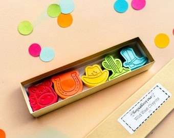 May include: A set of six colorful crayons shaped like cowboy-themed objects, including a horseshoe, a cactus, a cowboy hat, a boot, and a horse head. The crayons are packaged in a gold box with a label that reads "Kogelfreaky" and "Wild West Crayons".