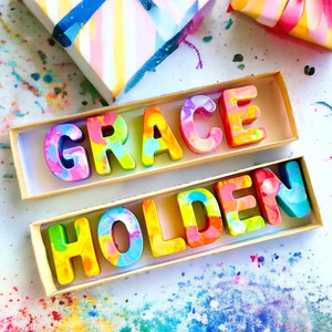 Op de afbeelding: Twee houten dozen met de namen "GRACE" en "HOLDEN" gespeld in kleurrijke, gemarmerde krijtletters. De dozen staan op een wit oppervlak met kleurrijke krijtjeskrullen eromheen.