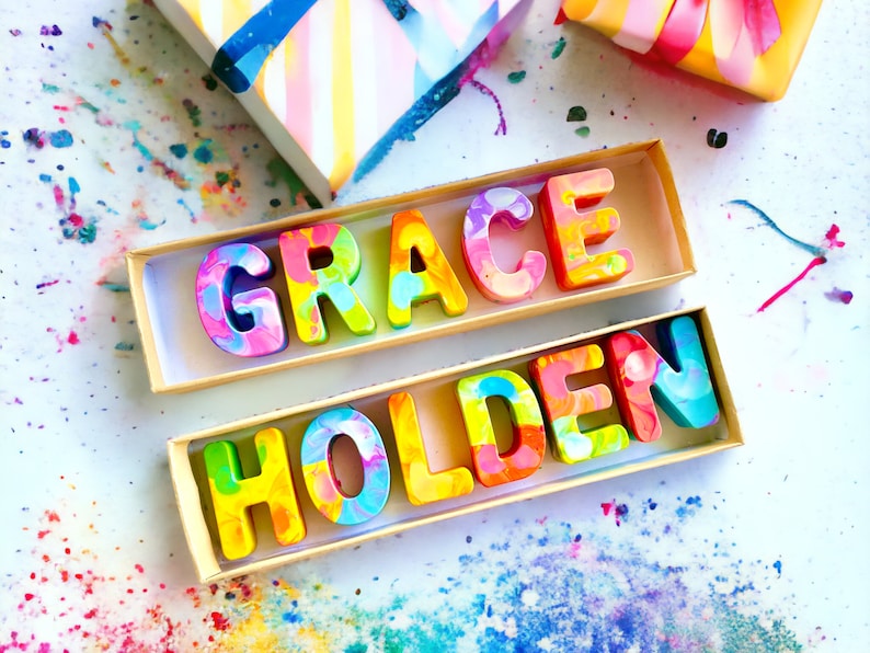 Op de afbeelding: Twee houten dozen met de namen "GRACE" en "HOLDEN" gespeld in kleurrijke, gemarmerde krijtletters. De dozen staan op een wit oppervlak met kleurrijke krijtjeskrullen eromheen.
