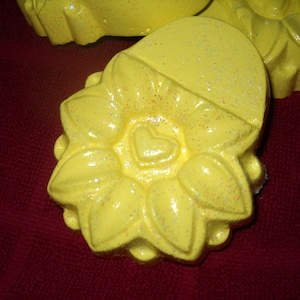 May include: Close-up of several yellow, sunflower-shaped soaps with a heart in the center. The soaps have a glittery finish and are set against a dark red background. The soaps are likely handmade and suitable for gifts.