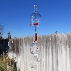 May include: A clear glass wind chime made from a repurposed wine bottle, with red accents and clear, curved acrylic rings. The wind chime hangs from a chain against a blue sky and a wooden fence.