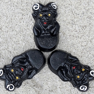 May include: Three black ceramic figurines with white and gold accents. Each figurine has a human-like face with a wide mouth and large eyes. The figurines are arranged in a triangular pattern.
