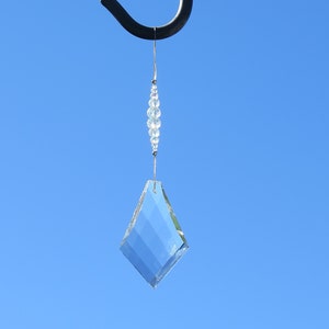 May include: A clear crystal prism hanging from a black metal hook against a bright blue sky. The prism is shaped like a diamond and has a string of clear beads above it.