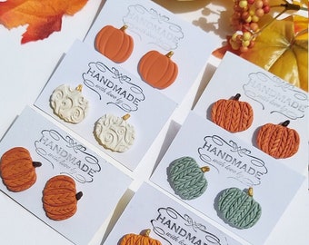 May include: A collection of handmade pumpkin earrings in various styles and colors, including orange, cream, and green. Each pair is displayed on a white card with the text "HANDMADE with love by". A decorative gourd and fall foliage are in the background.