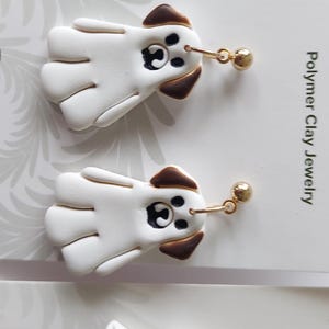 May include: A pair of white and brown dog-shaped dangle earrings made of polymer clay. The earrings feature a white body with brown ears and a black nose and eye detail, attached to gold-toned metal ball posts. The display card reads Blooming Fern Studio.
