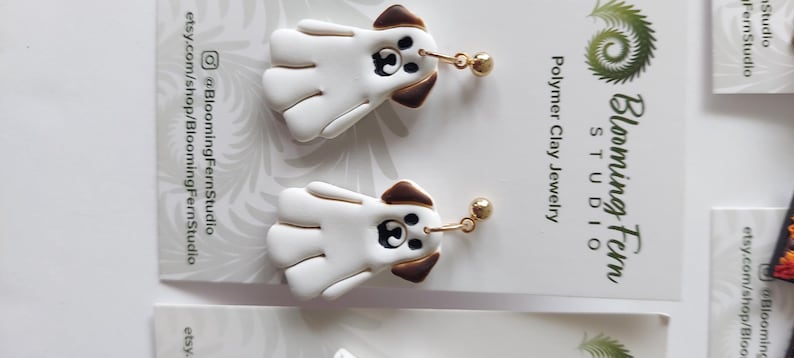 May include: A pair of white and brown dog-shaped dangle earrings made of polymer clay. The earrings feature a white body with brown ears and a black nose and eye detail, attached to gold-toned metal ball posts. The display card reads Blooming Fern Studio.