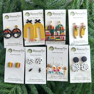 May include: Eight pairs of polymer clay earrings on white display cards. Designs include circular bookshelves, yellow pencil arches with black bows, rectangular bookshelves, pencil tips with flowers, small book stacks, music notes, paint palettes, and circles.