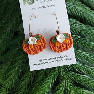 May include: Polymer clay earrings shaped like pumpkins with gold-colored hoops. The orange pumpkins have green leaves and white flower accents. The earrings are displayed on a white card with the Blooming Fern Studio logo and text.