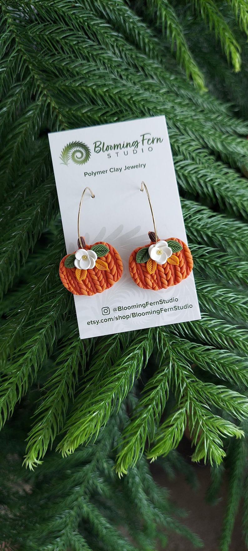 May include: Polymer clay earrings shaped like pumpkins with gold-colored hoops. The orange pumpkins have green leaves and white flower accents. The earrings are displayed on a white card with the Blooming Fern Studio logo and text.