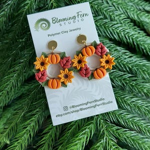 May include: Polymer clay earrings featuring a fall wreath design with pumpkins, sunflowers, and leaves. The earrings are in shades of orange, yellow, and red. The earrings are displayed on a white card with the Blooming Fern Studio logo and social media information.