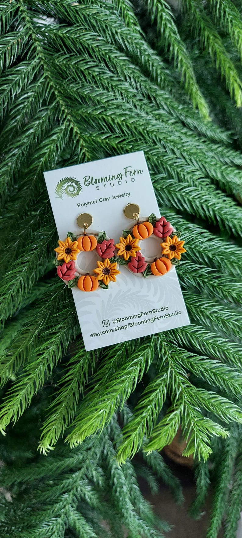 May include: Polymer clay earrings featuring a fall wreath design with pumpkins, sunflowers, and leaves. The earrings are in shades of orange, yellow, and red. The earrings are displayed on a white card with the Blooming Fern Studio logo and social media information.