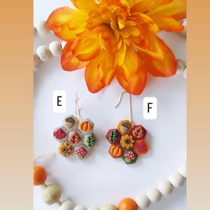 May include: Two pairs of handmade fall-themed earrings. Each earring features a honeycomb design with miniature pumpkins, acorns, leaves, and mushrooms in shades of orange, red, and brown. The earrings are displayed with a large orange flower and wooden beads.