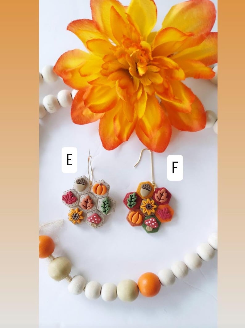 May include: Two pairs of handmade fall-themed earrings. Each earring features a honeycomb design with miniature pumpkins, acorns, leaves, and mushrooms in shades of orange, red, and brown. The earrings are displayed with a large orange flower and wooden beads.