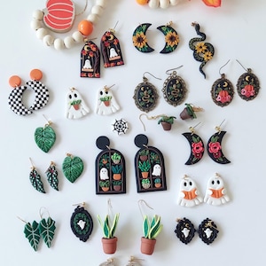 May include: A collection of polymer clay earrings featuring various designs including ghosts, potted plants, snakes, flowers, pumpkins, and checkered patterns. The set includes arched window frames, crescent moons, and oval frames with gold-toned hardware.