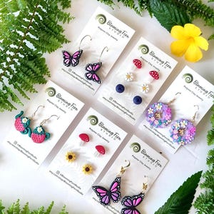 May include: A collection of polymer clay earrings featuring various designs, including butterflies, mushrooms, flowers, and teacups. The earrings are displayed on white cards with the text "Blooming Fern Studio" and shop information. The color palette includes pink, purple, red, yellow, and blue.