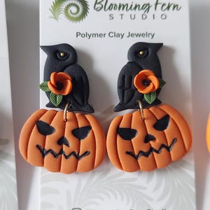 May include: A pair of polymer clay earrings on a white display card labeled Blooming Fern Studio. Each earring features a black crow perched atop an orange jack-o'-lantern with a carved face, accented by a small orange flower and green leaves.
