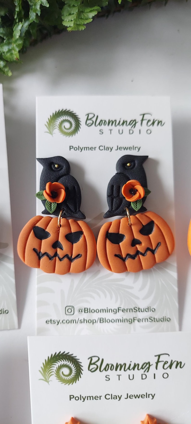 May include: A pair of polymer clay earrings on a white display card labeled Blooming Fern Studio. Each earring features a black crow perched atop an orange jack-o'-lantern with a carved face, accented by a small orange flower and green leaves.