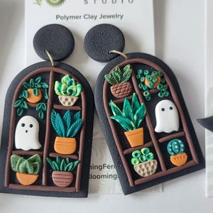 May include: A pair of black polymer clay earrings shaped like arched windows, featuring miniature potted plants and a white ghost figure. The earrings are attached to a white display card with the text Blooming Fern Studio Polymer Clay Jewelry.