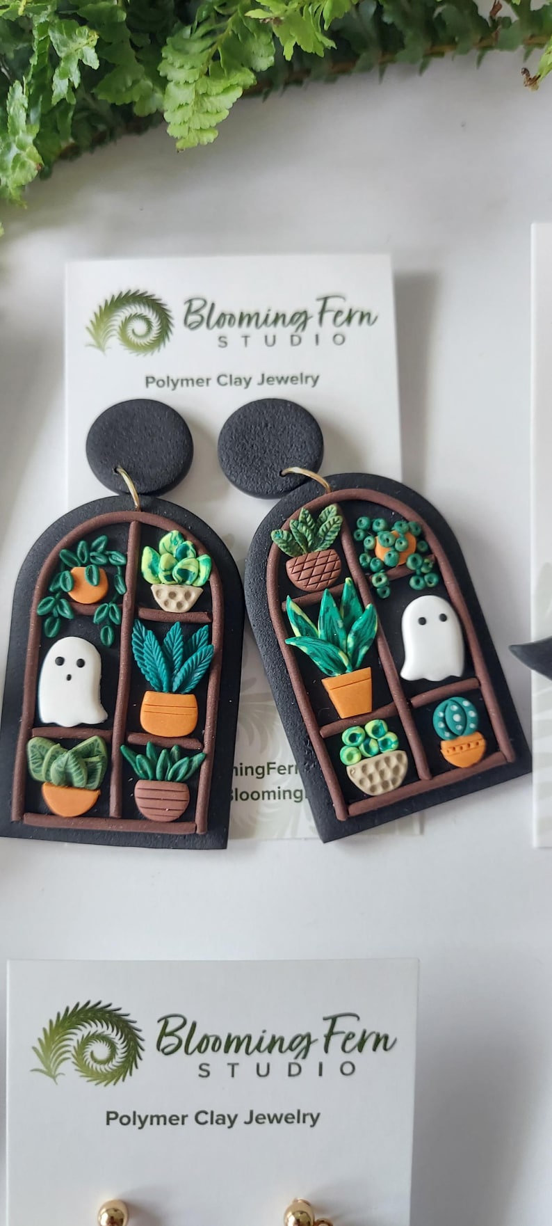 May include: A pair of black polymer clay earrings shaped like arched windows, featuring miniature potted plants and a white ghost figure. The earrings are attached to a white display card with the text Blooming Fern Studio Polymer Clay Jewelry.