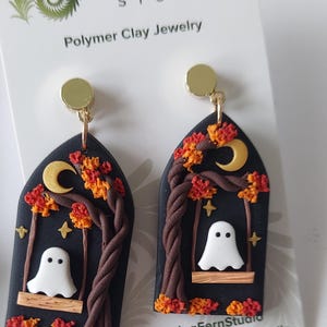 May include: A pair of arch-shaped polymer clay earrings featuring a black background with a white ghost sitting on a wooden swing. The design includes a brown twisted tree, orange and red autumn leaves, a yellow crescent moon, and small stars.