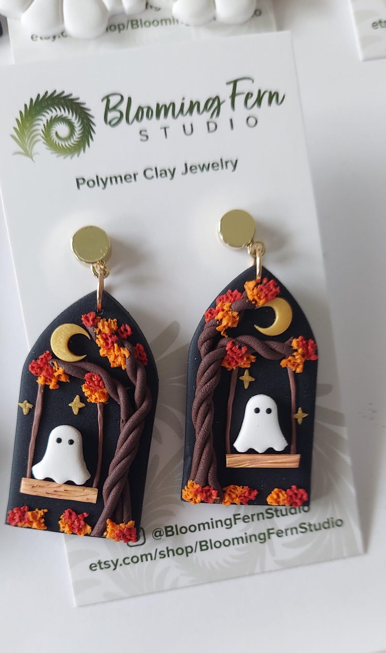 May include: A pair of arch-shaped polymer clay earrings featuring a black background with a white ghost sitting on a wooden swing. The design includes a brown twisted tree, orange and red autumn leaves, a yellow crescent moon, and small stars.
