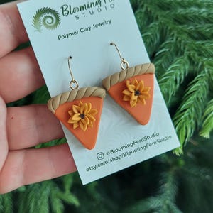 May include: A pair of polymer clay earrings shaped like pumpkin pie slices, each with a golden crust and a sunflower. The earrings are on a white card with the text "Blooming Fern Studio" and social media handles.