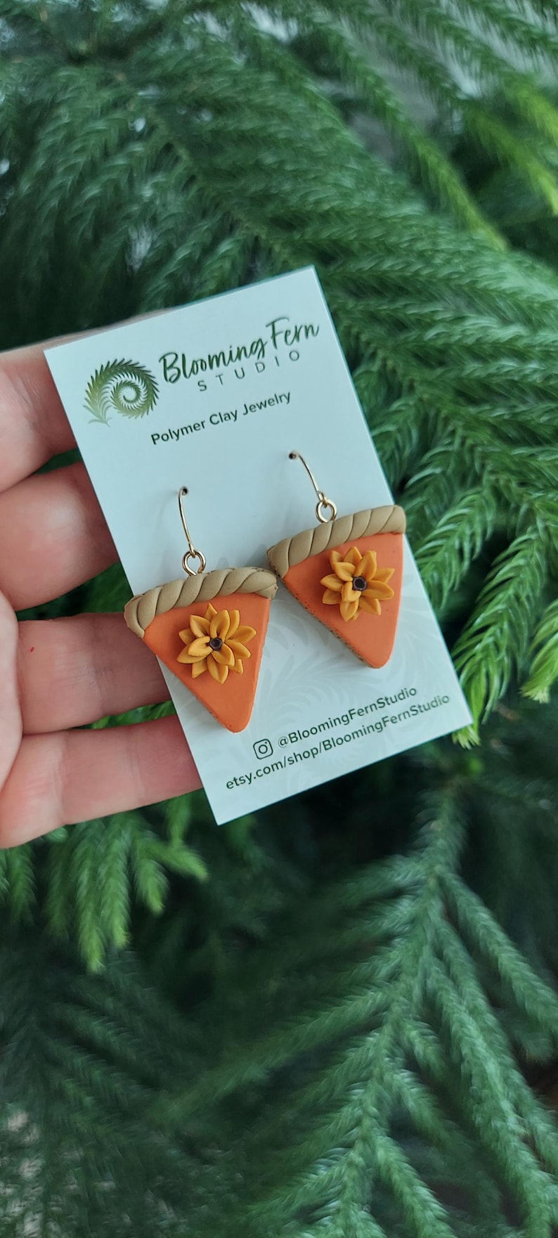 May include: A pair of polymer clay earrings shaped like pumpkin pie slices, each with a golden crust and a sunflower. The earrings are on a white card with the text "Blooming Fern Studio" and social media handles.