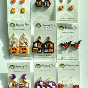 May include: A collection of polymer clay earrings with a Halloween theme. Designs include pumpkins, ghosts, candy corn, bats, books, and floral arrangements. Each pair is displayed on a white card with the Blooming Fern Studio logo.