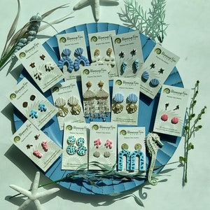 May include: A collection of polymer clay earrings displayed on a blue, circular tray. The earrings feature various ocean-themed designs, including seahorses, shells, and coral, in shades of blue, white, and gold. The cards read "Blaming Fern Studio".