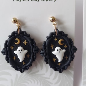 May include: A pair of dangle earrings featuring a white ghost, a gold crescent moon, and gold stars set against a black oval frame with a scalloped edge. The earrings are attached to gold-toned posts and displayed on a white card labeled Blooming Fern Studio Polymer Clay Jewelry.