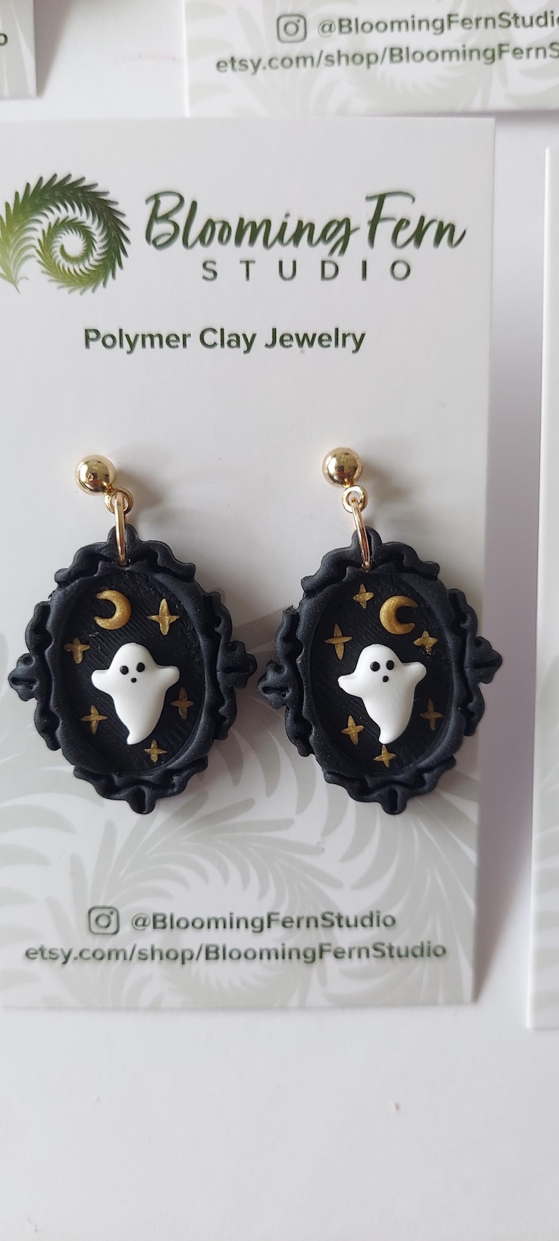 May include: A pair of dangle earrings featuring a white ghost, a gold crescent moon, and gold stars set against a black oval frame with a scalloped edge. The earrings are attached to gold-toned posts and displayed on a white card labeled Blooming Fern Studio Polymer Clay Jewelry.